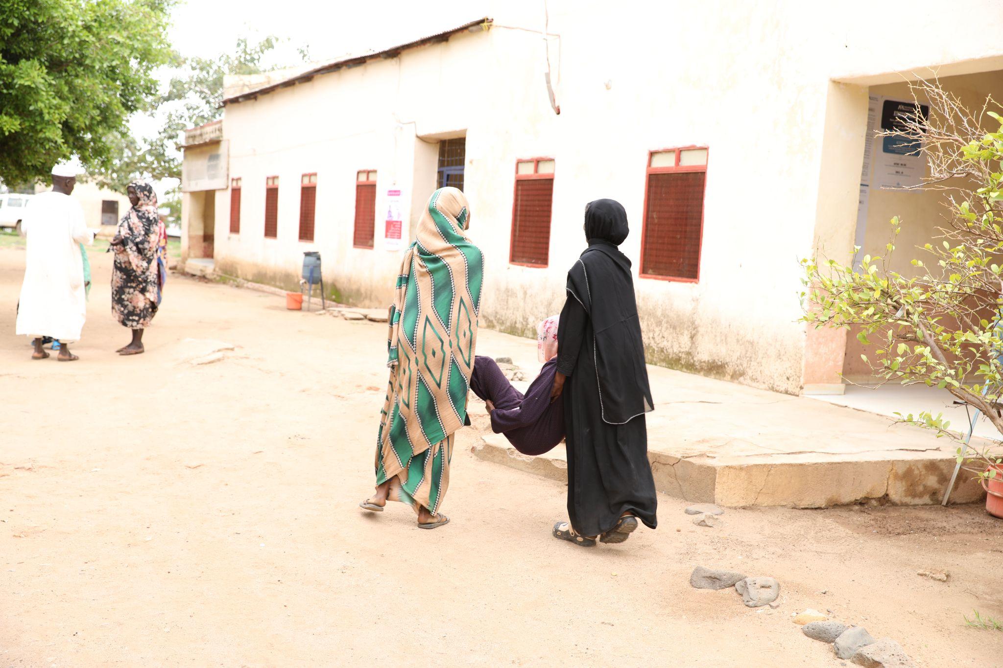 Two women carry another woman toward a clinic building in Sudan